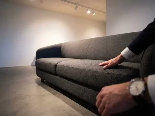Modern sofa in minimalist showroom with customer's hand reaching to touch realistic fabric texture under warm lighting.